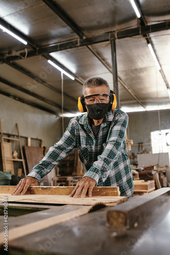 Experienced elderly carpenter in earmuffs cutting long wooden board with circular saw