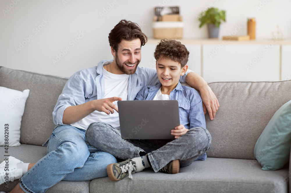 Caring daddy teaching son to use laptop or browsing internet together, sitting on comfy sofa in living room Caring daddy teaching son to use laptop or browsing internet together, sitting on comfy sofa in living room