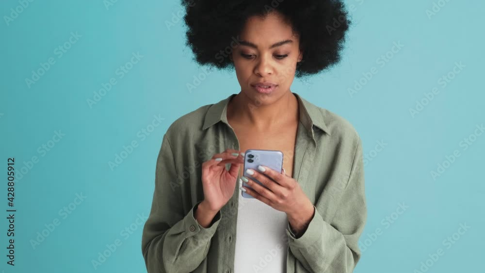 A black tense woman angrily answering someone by typing on the phone and standing in the studio A black tense woman angrily answering someone by typing on the phone and standing in the studio