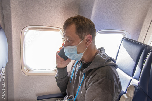 A 45-50-year-old man is sitting in the cabin of an airplane wearing a medical mask and talking on a mobile phone. Concept:violation of the rules of air travel, communication with loved ones during the
