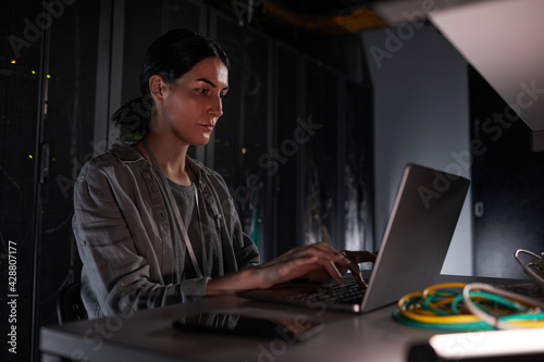 Portrait of female network engineer using laptop while sitting in dark server room, copy space