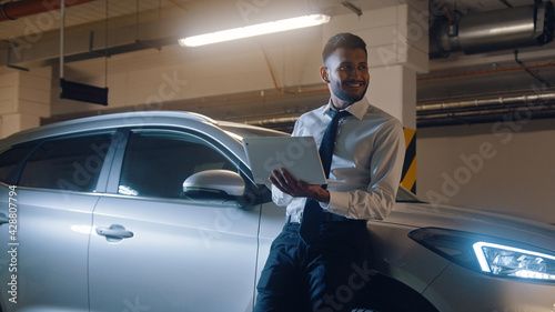 Happy smiling man leaning on silver car. Successful businessman holding laptop computer and looking away.