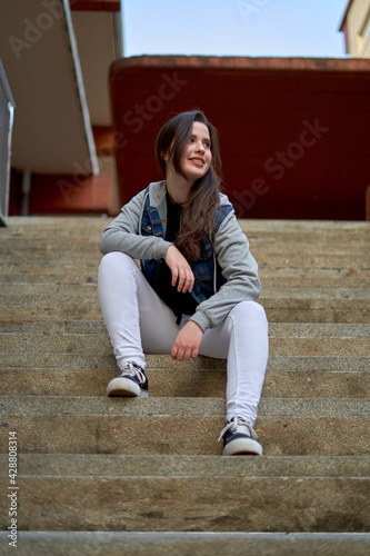 long-haired, fair-skinned teenager in denim jacket and white pants sitting on white stairs. Girl in urban style smiling and pensive. empowered young woman on the street © sacho