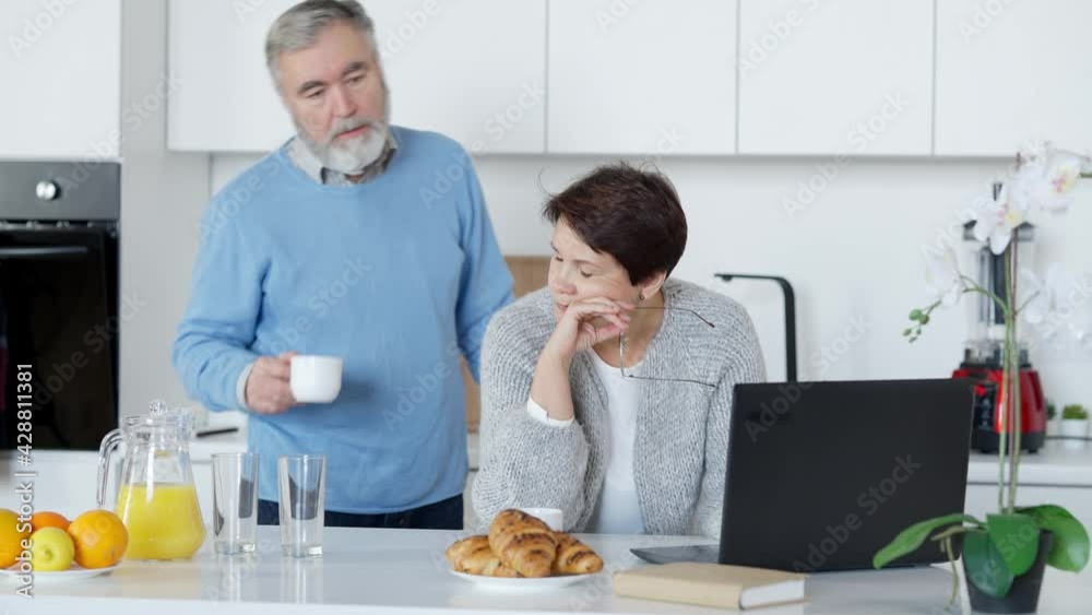 Loving senior man calming down stressed woman working online on laptop. Portrait of anxious Caucasian middle aged wife and caring husband in kitchen at home in the morning. Support and lifestyle Loving senior man calming down stressed woman working online on laptop. Portrait of anxious Caucasian middle aged wife and caring husband in kitchen at home in the morning. Support and lifestyle