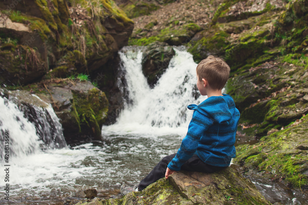 Kid sitting on the rock and looking at waterfall on the river in the rocks. Tourism with children concept. Kid sitting on the rock and looking at waterfall on the river in the rocks. Tourism with children concept.