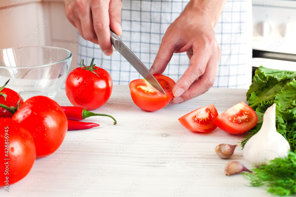 Man's hands cuts a ripe tomato with a knife on wooden white table. Process of making salad. Healthy food concept. Close-up. Man's hands cuts a ripe tomato with a knife on wooden white table. Process of making salad. Healthy food concept. Close-up.