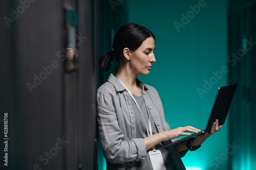 Side view portrait of female data engineer holding laptop while working with supercomputer in server room, copy space photo