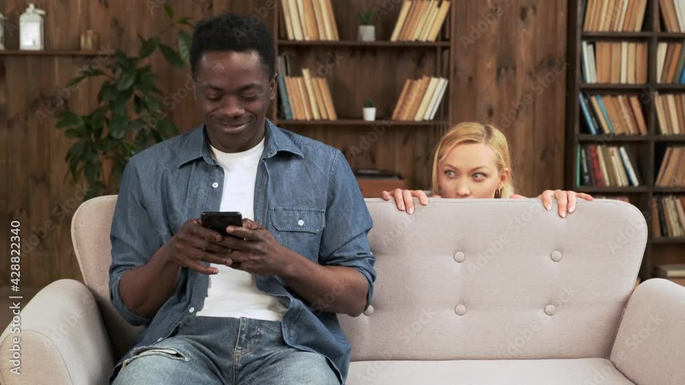 Cheater man dating online with a smart phone and girlfriend is spying sitting on a sofa at home. Mixed race ethnicity couple