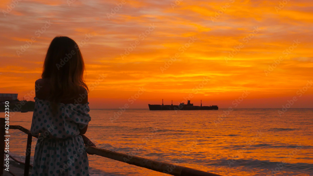 Woman with blonde and long hair, in a dress with bare shoulders looks at the sunrise and the ship at sea on the coast. 