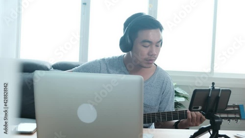 Musician Asian male playing acoustic guitar singing song learning practicing carefree activity occupation sitting on sofa using laptop mobile recording live stream social media entertaining people