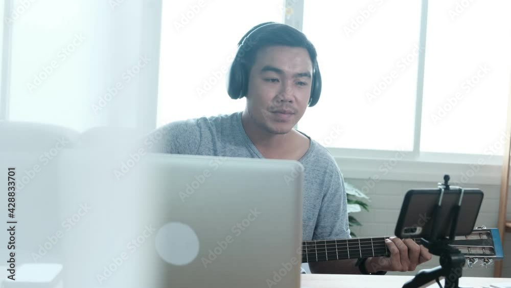 Musician Asian male playing acoustic guitar singing song learning practicing carefree activity occupation sitting on sofa using laptop mobile recording live stream social media entertaining people Musician Asian male playing acoustic guitar singing song learning practicing carefree activity occupation sitting on sofa using laptop mobile recording live stream social media entertaining people