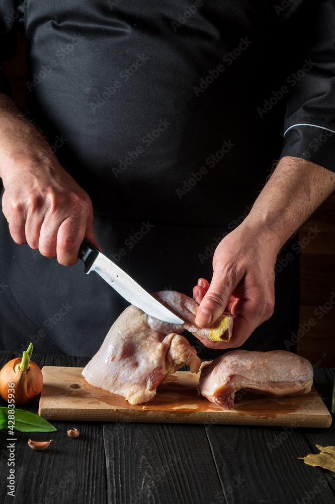 Preparing for cooking chicken legs in the restaurant kitchen. Chef cuts a chicken leg with a knife on cutting board. Grilling chicken