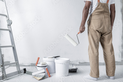 a man paints a white wall with a roller. Repair of the interior. Young male decorator painting a wall in the empty room, concept builder or painter in helmet with paint roller over the empty room