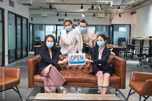 Group of asian business people wearing face mask sitting and holding reopen and keep social distancing paper in the office