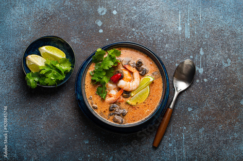 Traditional spicy Thai soup Tom Yum with shrimp and seafood in ceramic bowl on blue stone rustic background from above, classic dish of cuisine of Thailand 
