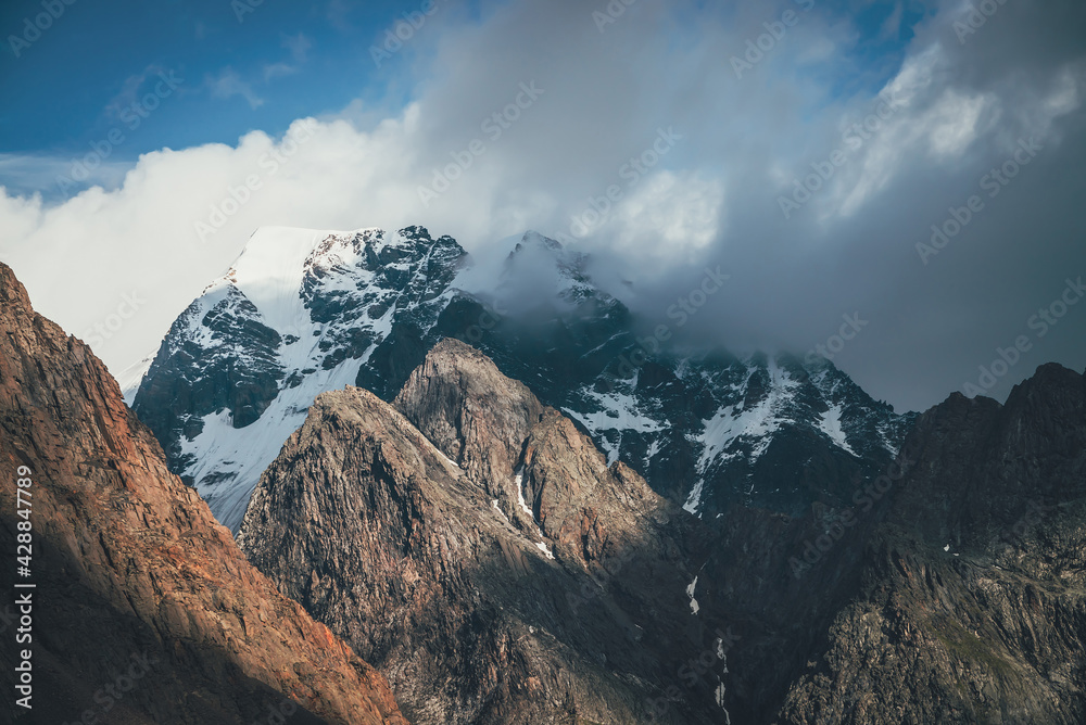 Obraz premium Scenic landscape with great rocks and snowy mountains in sunlight in low clouds. Wonderful view to mountain top with snow in sunshine in cloudy sky. Awesome scenery with sunlit snow-white pinnacle.