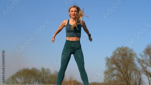 Athletic girl in green sportswear doing warm-up exercises before training outdoors. Woman athlete are doing sports exercise jumping on blue sky background.