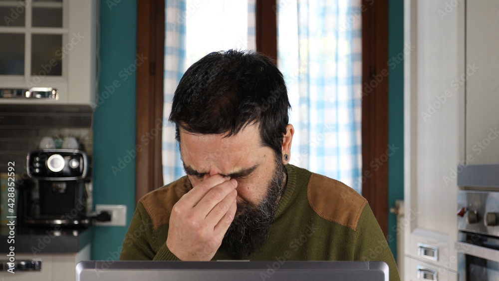 Attractive bearded man working on computer. Sitting at desk in home typing on laptop tired rubbing nose and eyes feeling fatigue and headache. Stress and frustrated concept