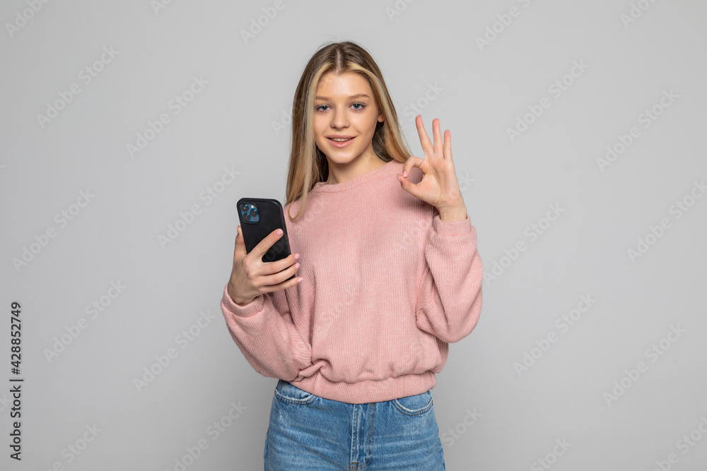 Fototapeta premium Attractive woman showing ok gesture while holding blank screen mobile phone over gray background