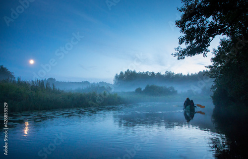 Hikers rowing an canoe in the summer night river, surrounded by fog clad floodplain and under full moon in the sky on the Halliste river in Soomaa NP 