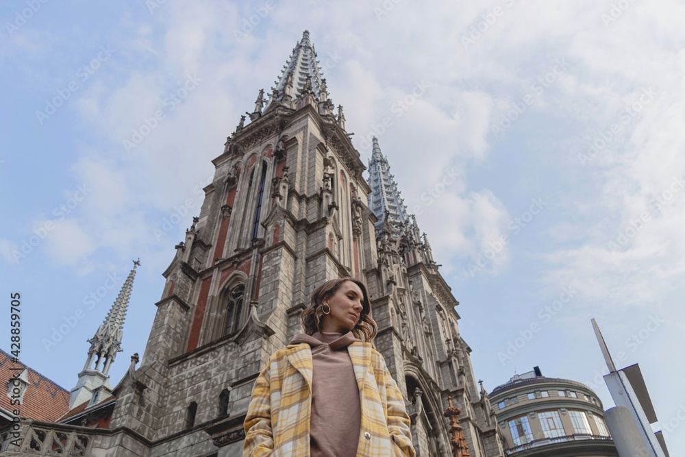Fototapeta premium Happy millenial woman in a yellow coat in European city near historic building. Local travel concept.