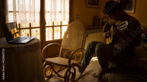 man ridiculously learning to play guitar using a laptop while sitting on a bed at home