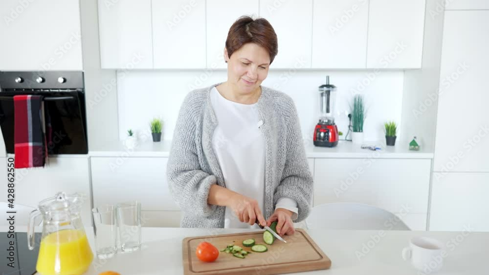 Beautiful middle aged Caucasian woman slicing cucumber and tasting delicious vegetable in kitchen at home. Portrait of positive confident lady cooking healthful vegan salad indoors smiling and Beautiful middle aged Caucasian woman slicing cucumber and tasting delicious vegetable in kitchen at home. Portrait of positive confident lady cooking healthful vegan salad indoors smiling and