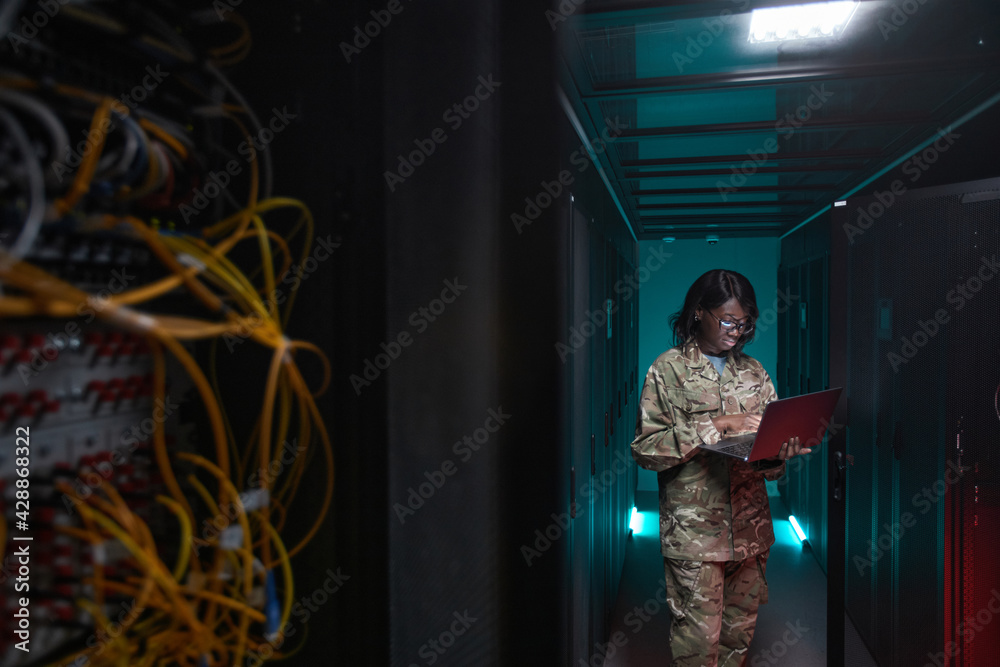 © Seventyfour - Portrait of young African-American woman wearing military uniform using laptop while standing in server room, copy space