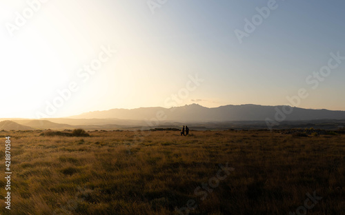Dos personas pasean durante el atardecer por el campo, con unas grandes montañas al fondo y hierba seca photo