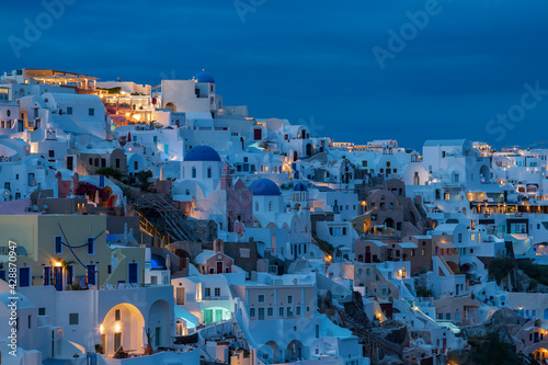 Evening view from the viewpoint of Oia town on Santorini in Greece after sunset. Beautiful clouds taken long time. The background is the sky at the blue hour. Lights are shining in white houses. © Roman Bjuty