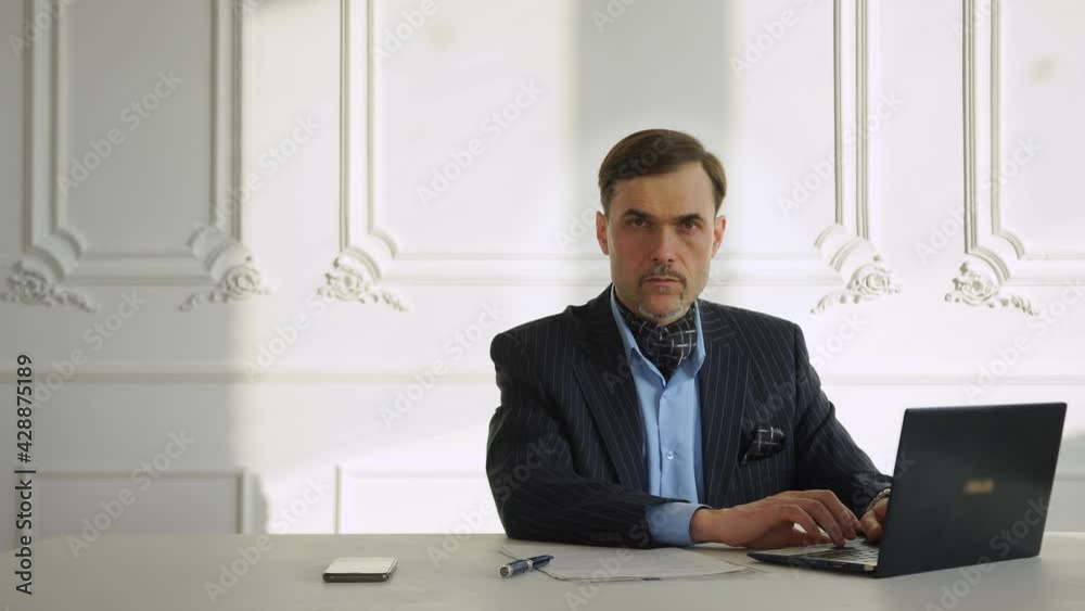 Businessman, lawyer, financier, trader, entrepreneur works, prints, studies the market, reads laptop blue suit shirt sitting table in large white room. Place to insert text. Looks into the camera