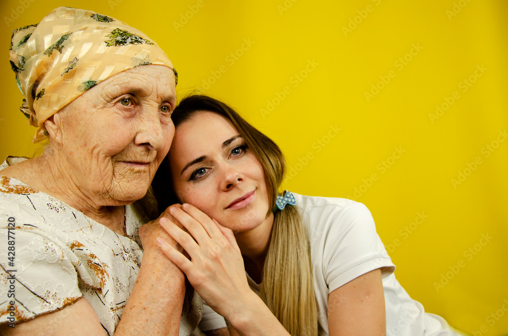 The granddaughter leaned her head on her grandmother's shoulder. Old hand of grandmother in young hands. Grandmother and granddaughter on a yellow background. Two generations of women. The granddaughter leaned her head on her grandmother's shoulder. Old hand of grandmother in young hands. Grandmother and granddaughter on a yellow background. Two generations of women.