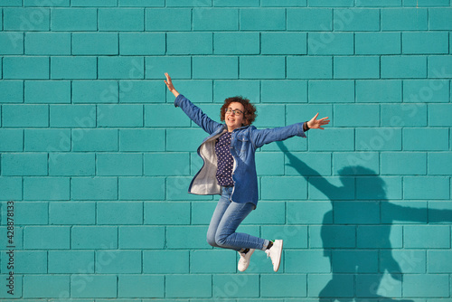 Front view of a happy woman jumping against a bright blue wall on a sunny day