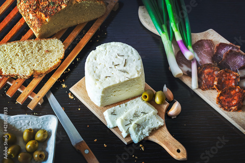 Goat cheese sliced on a wooden plate with freshly baked bread with green onions, smoked ham, sausage, olives on a dark wooden dining table.Top view