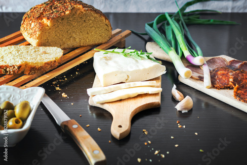 Cheese sliced on a wooden plate with freshly baked bread with green onions, smoked ham, sausage, olives on a dark wooden dining table.Top view
