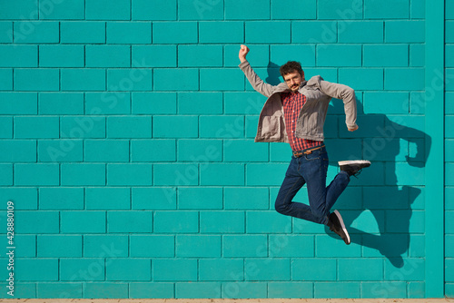 Front view of a happy man jumping against a blue bright wall in a sunny day