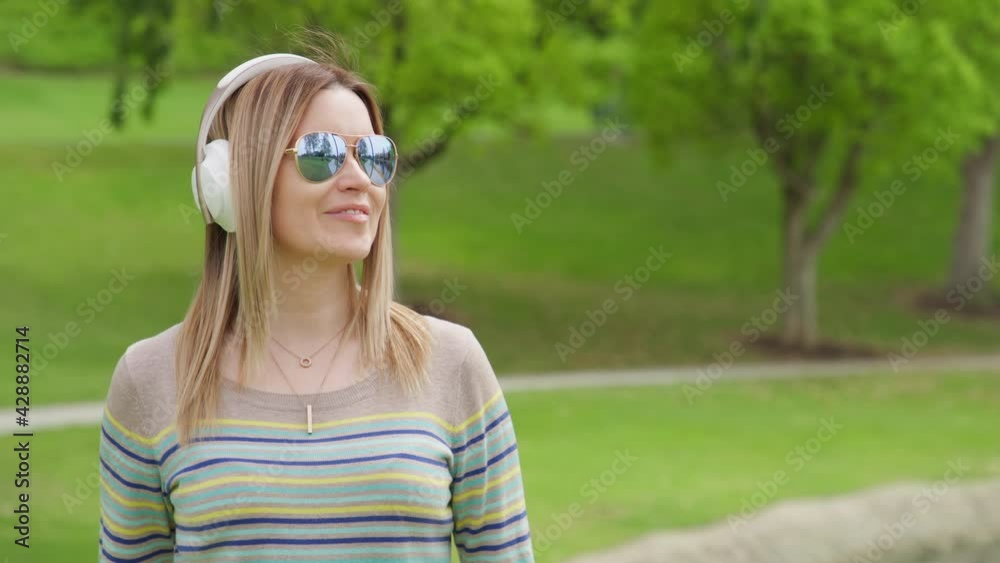 Portrait of young caucasian blonde woman with headphones listening to music, dreaming and moving to the beat in green city park. Copy space on green background. RED helium weapon footage 8K