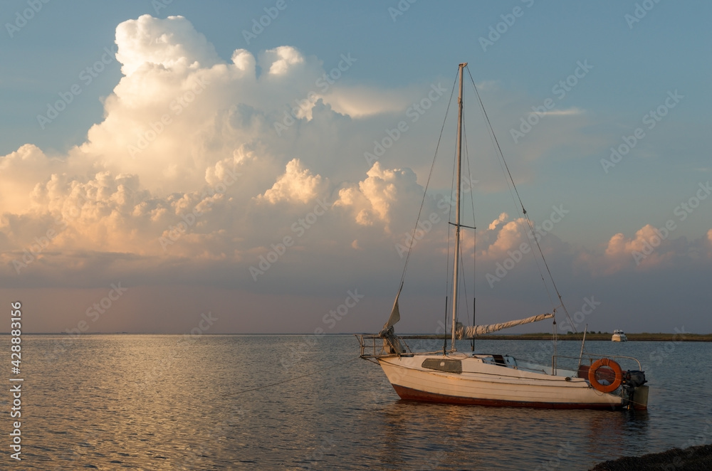 Fototapeta premium Small white yacht docked by the coast against an evening sky with big cloud. Sunset over Black sea, summer time. Family vacation or travel by sea on own yacht.