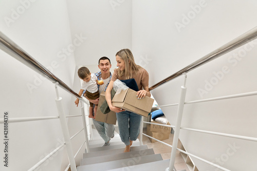 Happy family couple new home owners real estate buyers or renters with toddler kid child son carrying boxes going up stairs on moving day arriving into new apartment. Relocation, mortgage, removals. photo