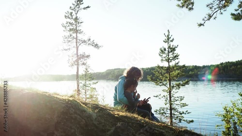 Happy curly hair mother with little daughter sitting by lake during sunset on spring camping vacation and browsing internet on mobile phone device watching video. Concept of nature quality family time