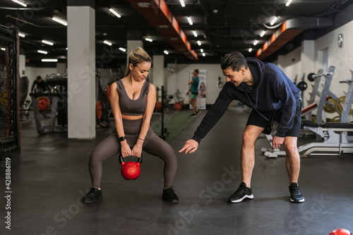 Fitness couple in sportswear squatting with weights and doing squats in the gym. A personal trainer in the gym corrects the squats of a young athlete