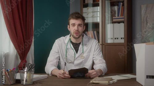 Confident young American doctor reading x-ray picture, concentrated facial expression, treating patient online using video chat internet connection on laptop computer photo