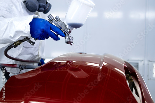 Fototapeta Naklejka Na Ścianę i Meble -  Car painter in protective clothes and mask painting automobile bumper with metallic paint and varnish in chamber workshop.