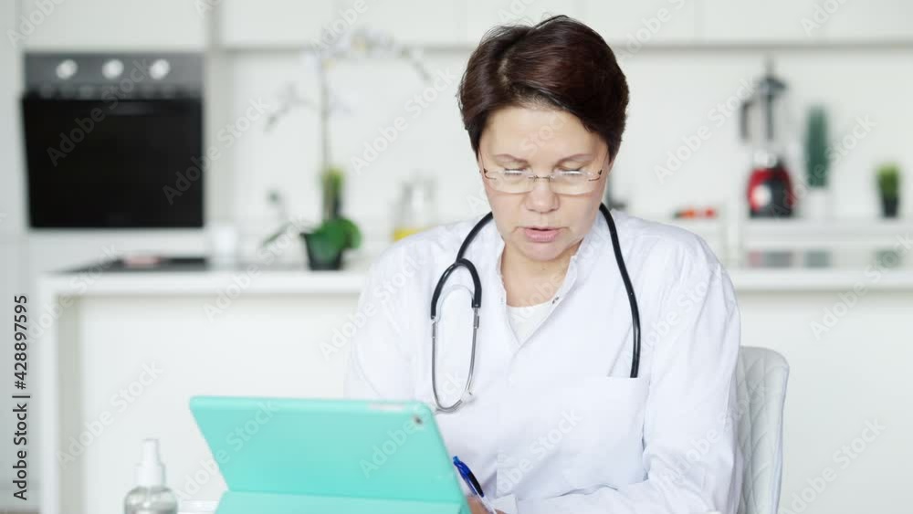 Concentrated professional doctor consulting patient online talking and writing symptoms. Portrait of serious middle aged Caucasian woman working from home on coronavirus pandemic. New normal Concentrated professional doctor consulting patient online talking and writing symptoms. Portrait of serious middle aged Caucasian woman working from home on coronavirus pandemic. New normal