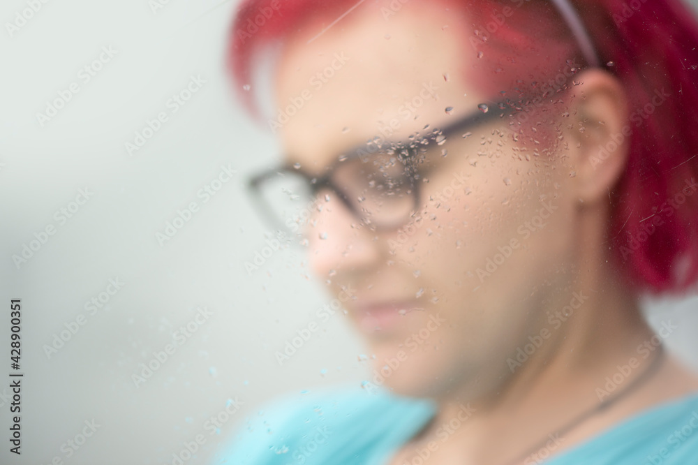 Obraz premium Frau mit roten Haaren und Brille, Nahaufnahme durch Fensterscheibe mit Wassertropfen, unscharfer Hintergrund