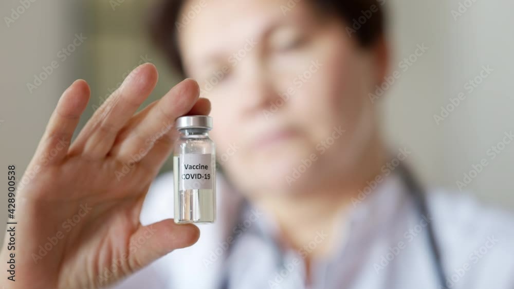 Close-up of coronavirus vaccine jar in female Caucasian hand with blurred face of serious middle aged woman at background. Professional doctor scientist showing Covid-19 treatment indoors Close-up of coronavirus vaccine jar in female Caucasian hand with blurred face of serious middle aged woman at background. Professional doctor scientist showing Covid-19 treatment indoors