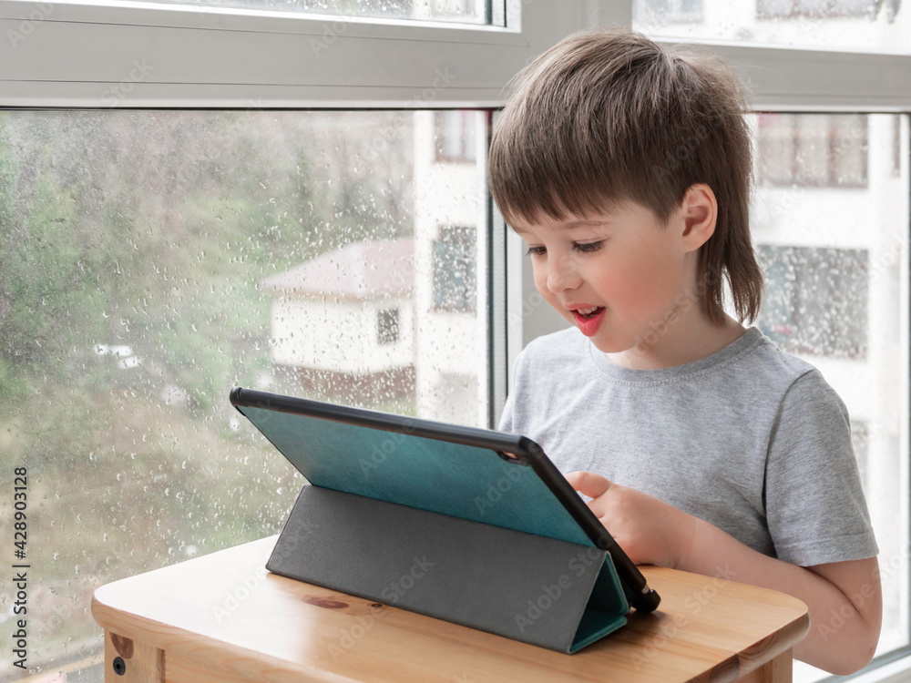 Curious boy watch cartoons on digital tablet. Kid uses electronic device. Indoor leisure for children while it's raining outside. Curious boy watch cartoons on digital tablet. Kid uses electronic device. Indoor leisure for children while it's raining outside.