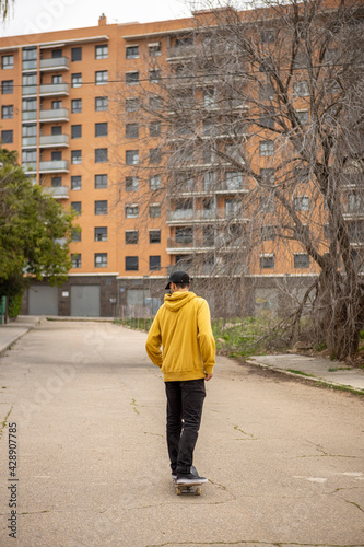 Male in a yellow hoodie and black cap on the skateboard in the background of a high rise building