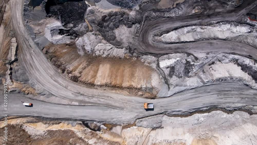 Brown coal mining section. View from above. A yellow dump truck drives along a dirt road among a coal mine. Coal mining. Heavy mining equipment. Brown coal mining section. View from above. A yellow dump truck drives along a dirt road among a coal mine. Coal mining. Heavy mining equipment.