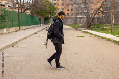 Young male with a black coat and cap walking on the street with a skateboard